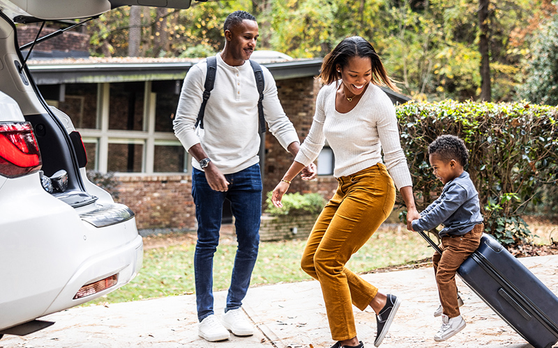 Couple with child sitting on suitcase in the driveway near trunk of car